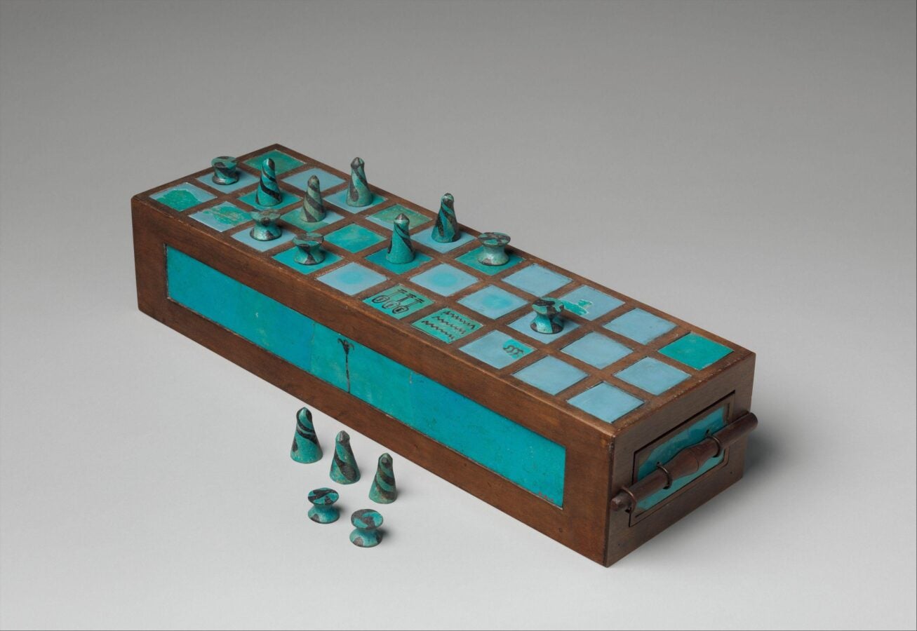 A rectangular ancient Egyptian game board with turquoise and brown squares, featuring small cone-shaped and peg-like playing pieces arranged on top and beside it.